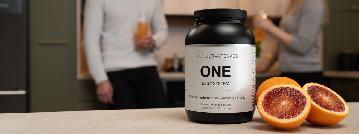 Ultimate Labs ONE Daily System -ravintolisäpurkki, veriappelsiinit ja kaksi henkilöä pitelemässä juomia taustalla keittiössä